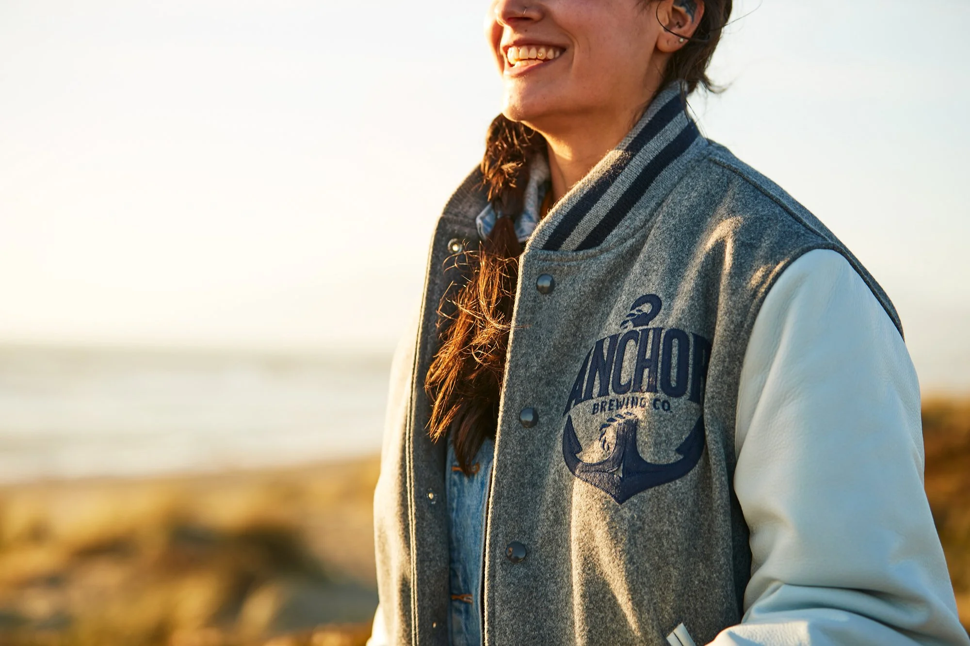 A person smiling outdoors wearing a gray and white varsity jacket with “ANCHOR BREWING CO” written on it, sunlight illuminating their face and hair, with a blurred natural background.