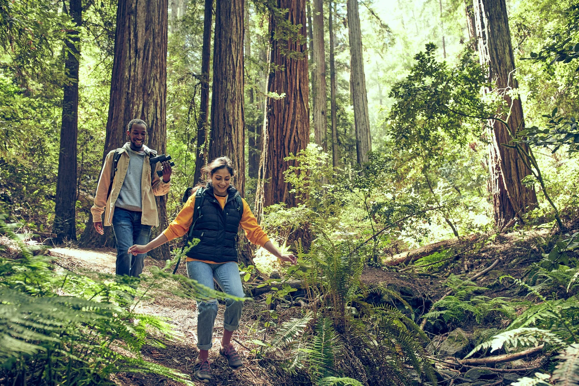 Two smiling women hiking through a forest with tall trees and lush greenery, one holding binoculars.