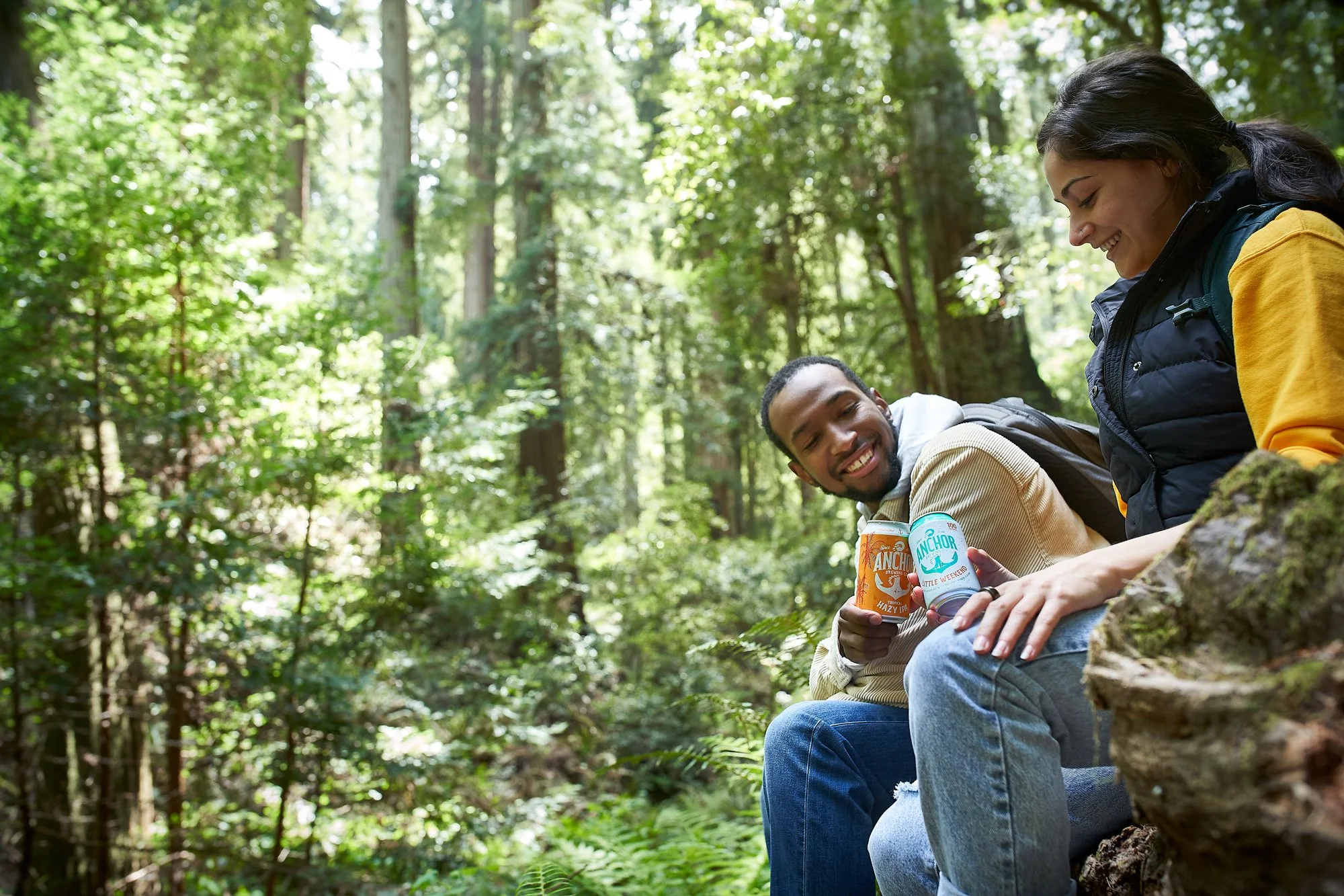 Two friends sitting on a fallen log in a lush green forest, smiling and holding cans of Anchor Brewing beer.
