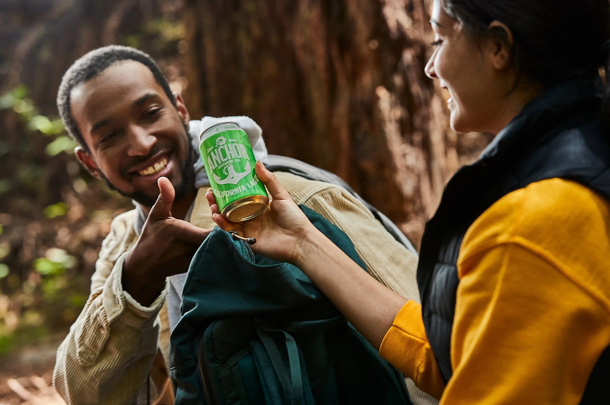 Two people smiling and interacting outdoors in a wooded area; one is holding a green can of Anchor California Lager, and the other person is reaching for it.