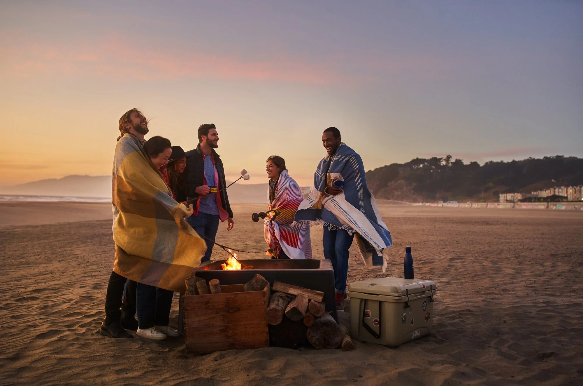 Group of people gathered around a fire on the beach during sunset, with some wrapped in blankets, smiling and enjoying each other's company.