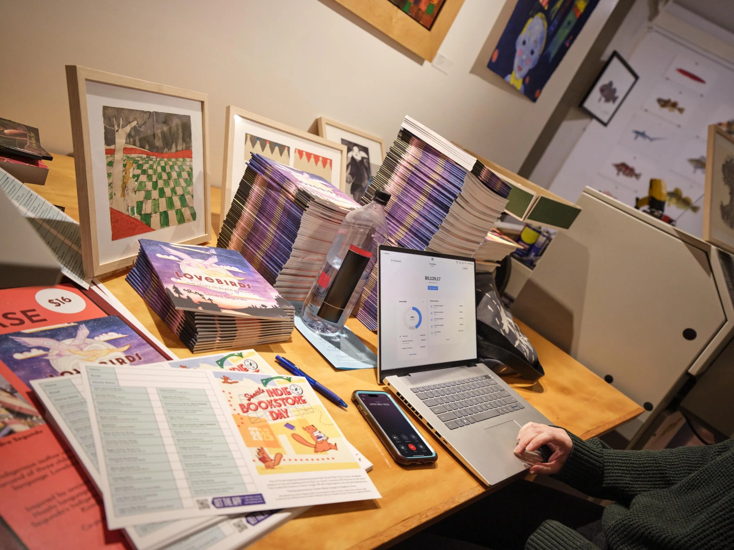 A table displays colorful books, illustrated posters, flyers for a bookstore day, a laptop, smartphone, and a water bottle, in front of a wall with artwork.