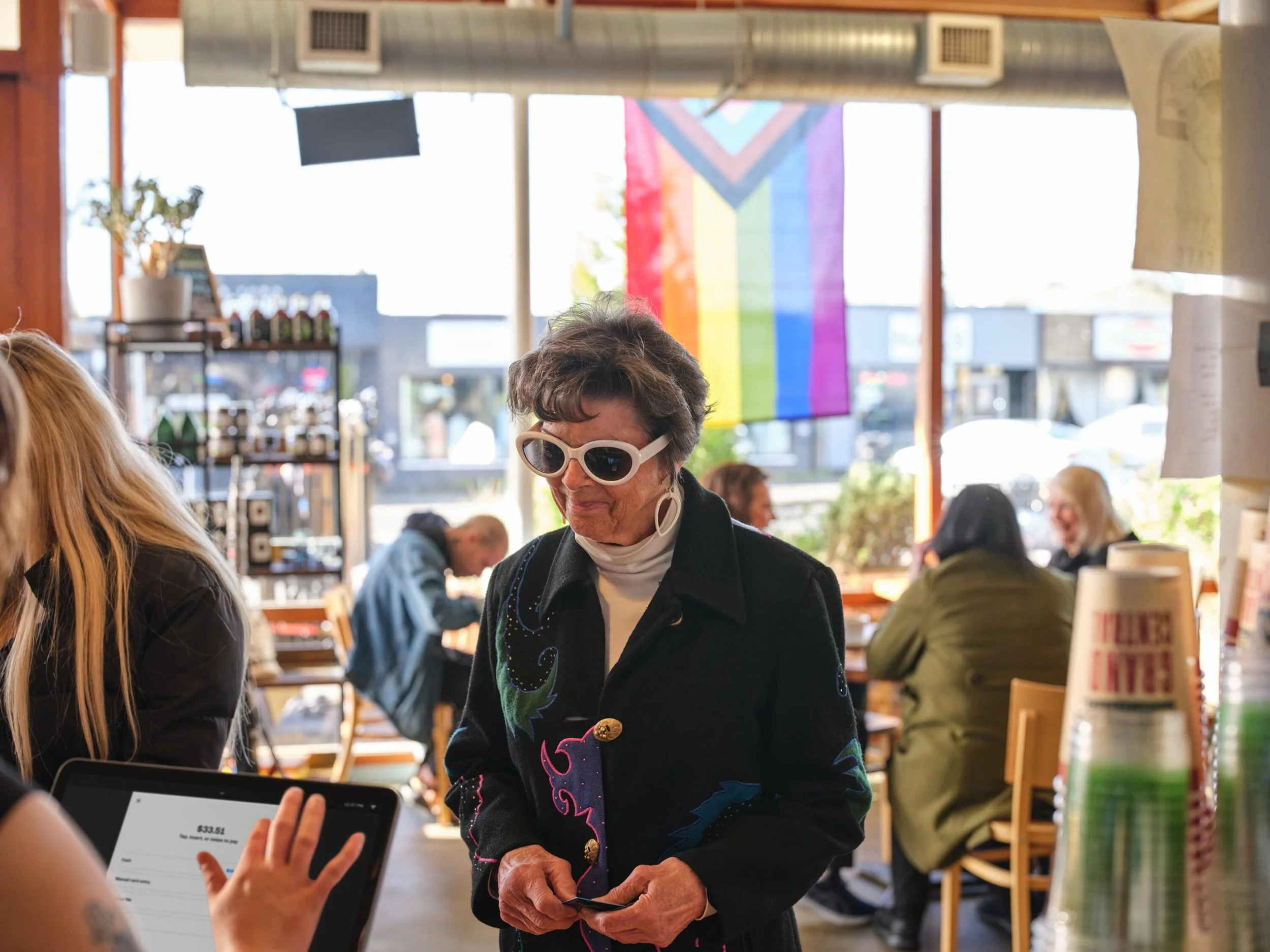 An elderly woman wearing sunglasses and a black embroidered jacket is in conversation with a person holding a smartphone inside a restaurant. The background features large windows, a rainbow pride flag, and several patrons dining.