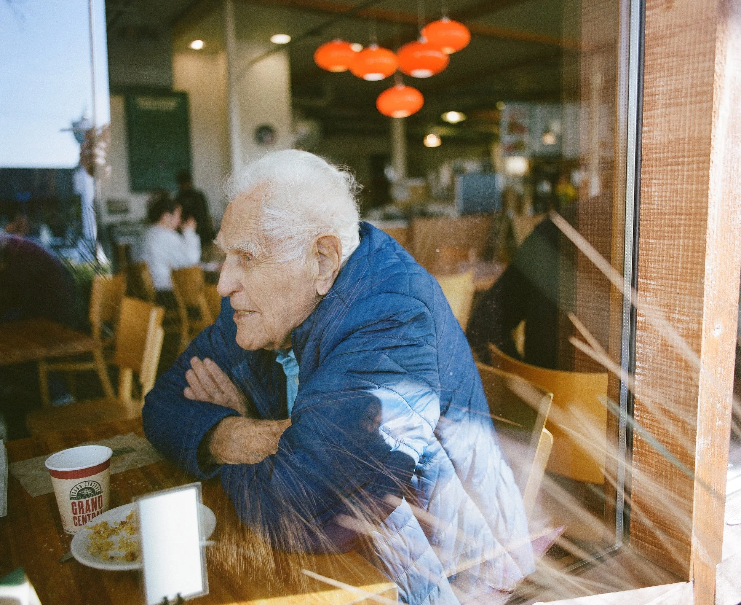 An elderly woman with white hair and glasses, wearing a blue jacket, sitting at a table inside a restaurant, viewed through a window with a reflective surface. Coffee cups and a plate with food are on the table.