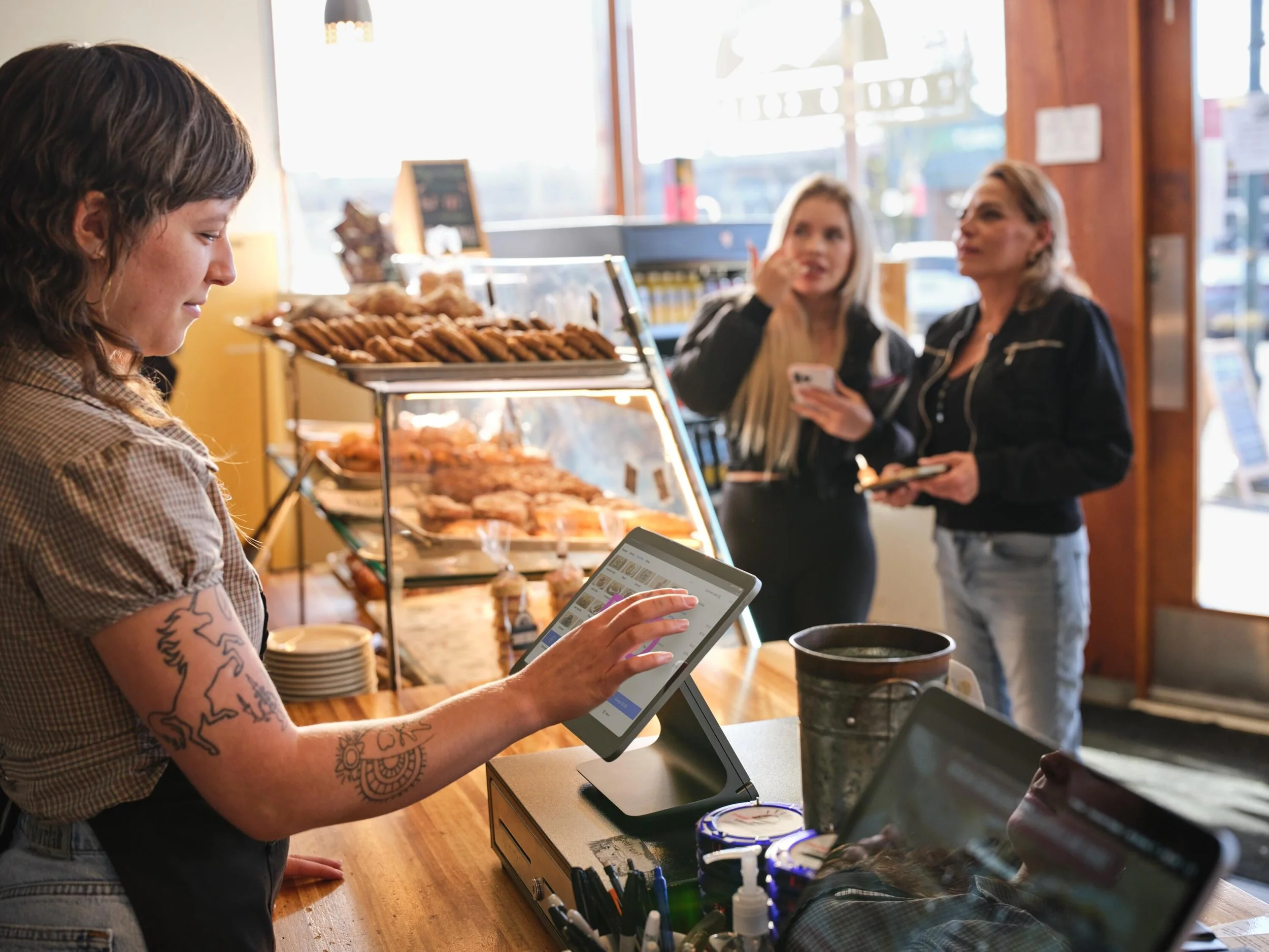 A woman with tattoos on her arm is using a tablet at a counter in a bakery or cafe. Behind her, two women are talking near the display of baked goods, with one holding a phone.