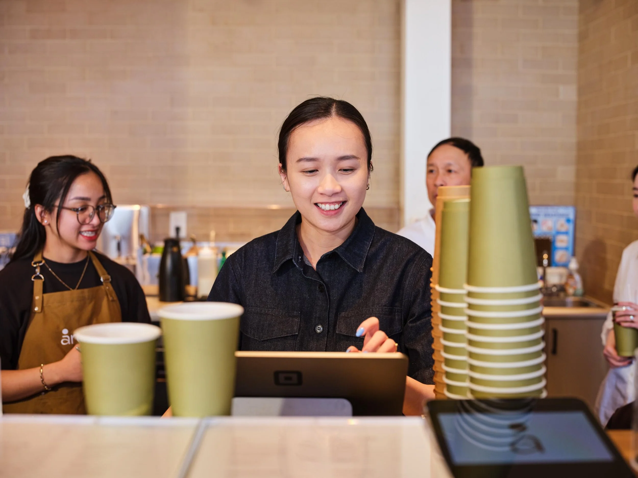 Three women working at a coffee shop counter, with one woman in the center smiling and looking down at a laptop. The woman on the left is wearing glasses and a brown apron, and the background features a brick wall and coffee cups.