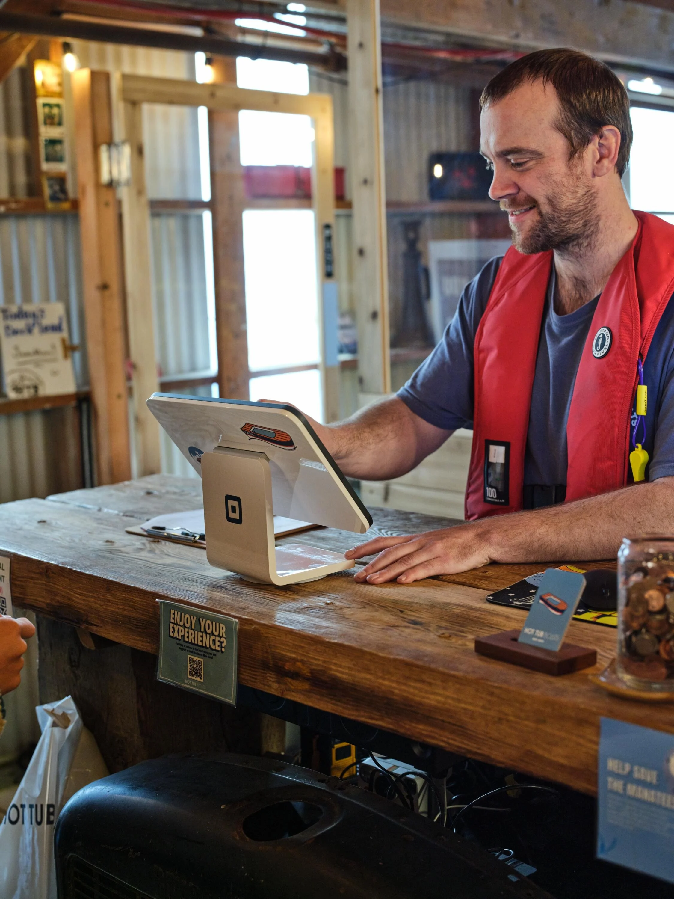 A man with short dark hair and a beard wearing a red vest and t-shirt is smiling while using a touchscreen point of sale register at a rustic wooden counter in a store. Shelving with products and bright windows are in the background.