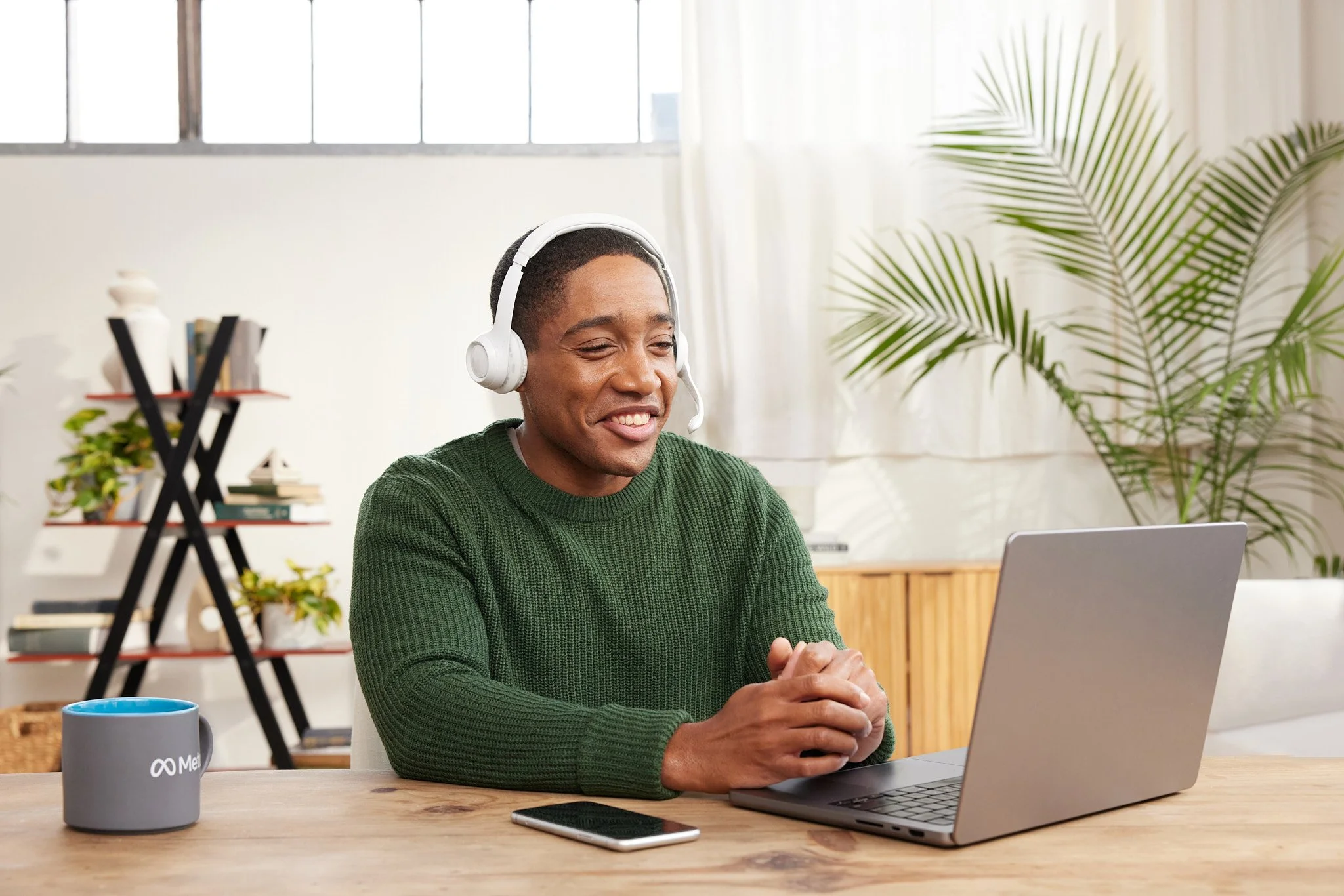 Man wearing a green sweater and white headset using a laptop at a wooden table in a bright room, with a large plant and bookshelf in the background, and a mug on the table.