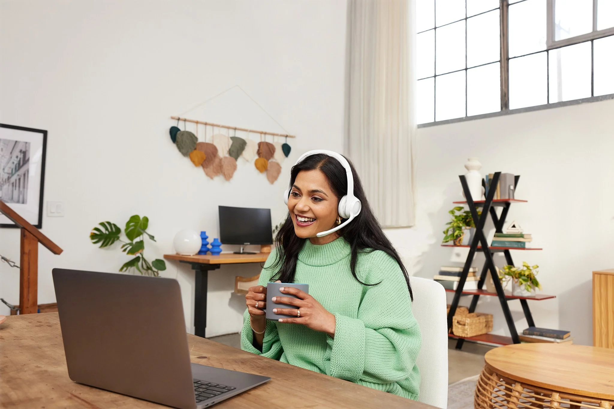 Woman wearing headphones sitting at a table with a laptop, holding a mug, in a bright, modern room.