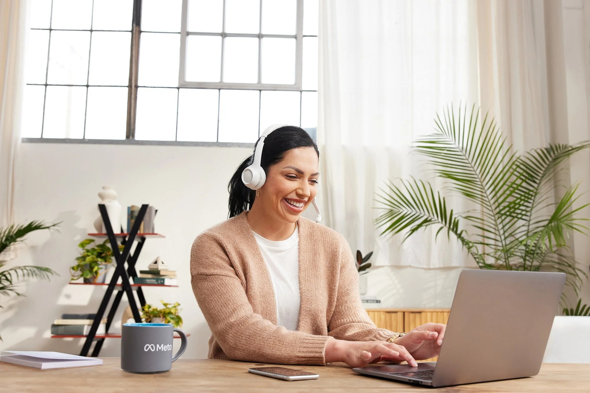 A woman with dark hair smiling while working on a laptop in a bright room with large windows and plants.