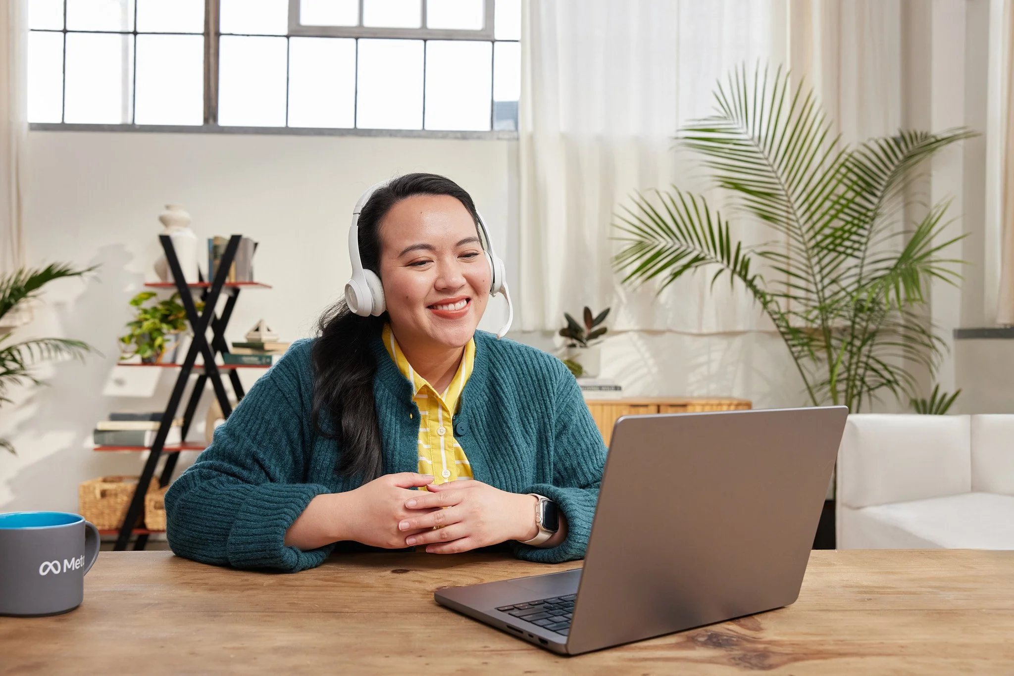A young woman with long dark hair wearing a blue sweater and yellow shirt, smiling as she looks at her tablet while sitting at a wooden table with a laptop and a mug in a cozy, well-lit room with plants and large windows.