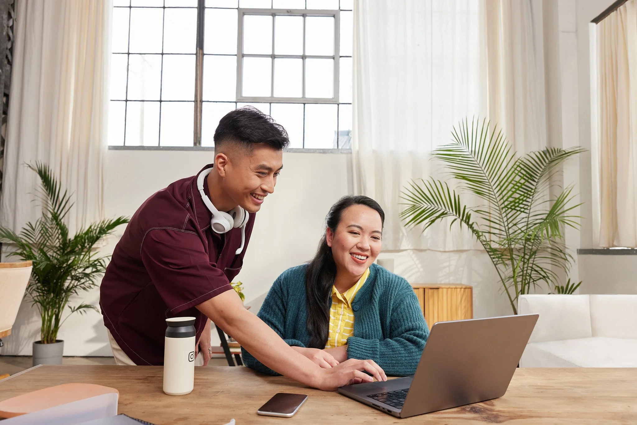 A young man with headphones around his neck helps a woman at a laptop in a well-lit room with large windows, white curtains, and potted plants on the windowsill.