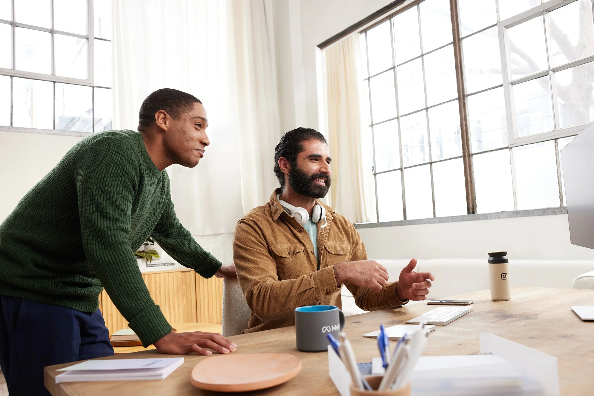 Two men working together at a desk in a bright office, with large windows and various office supplies around.