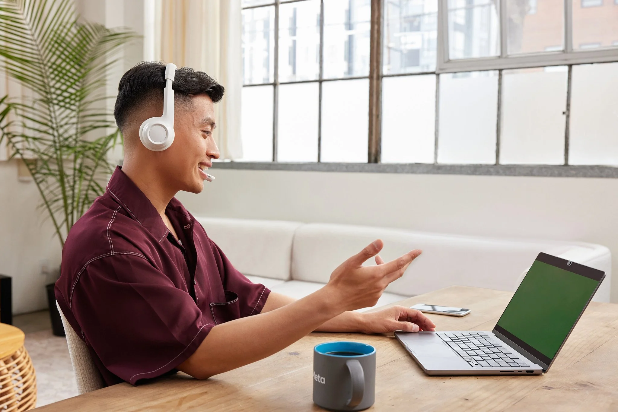 A young man with dark hair wearing a burgundy shirt, sitting at a wooden table, smiling and wearing a white headset. He is gesturing with his right hand while looking at a laptop with a green screen. On the table, there is a grey coffee mug with blue inside and a smartphone. The background features large windows and a potted plant.