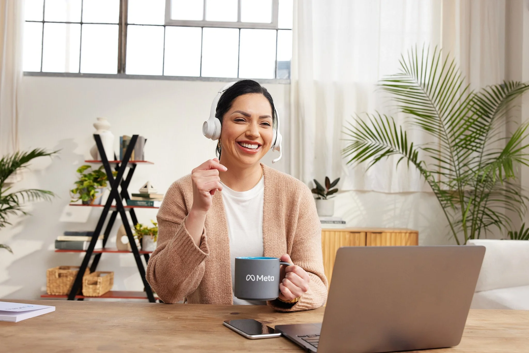 A woman with dark hair, wearing a beige cardigan and white shirt, smiling and holding a gray mug with Meta logo on it, sitting at a wooden table with an open laptop, smartphone, and papers. She is wearing a headset with a microphone. The background f