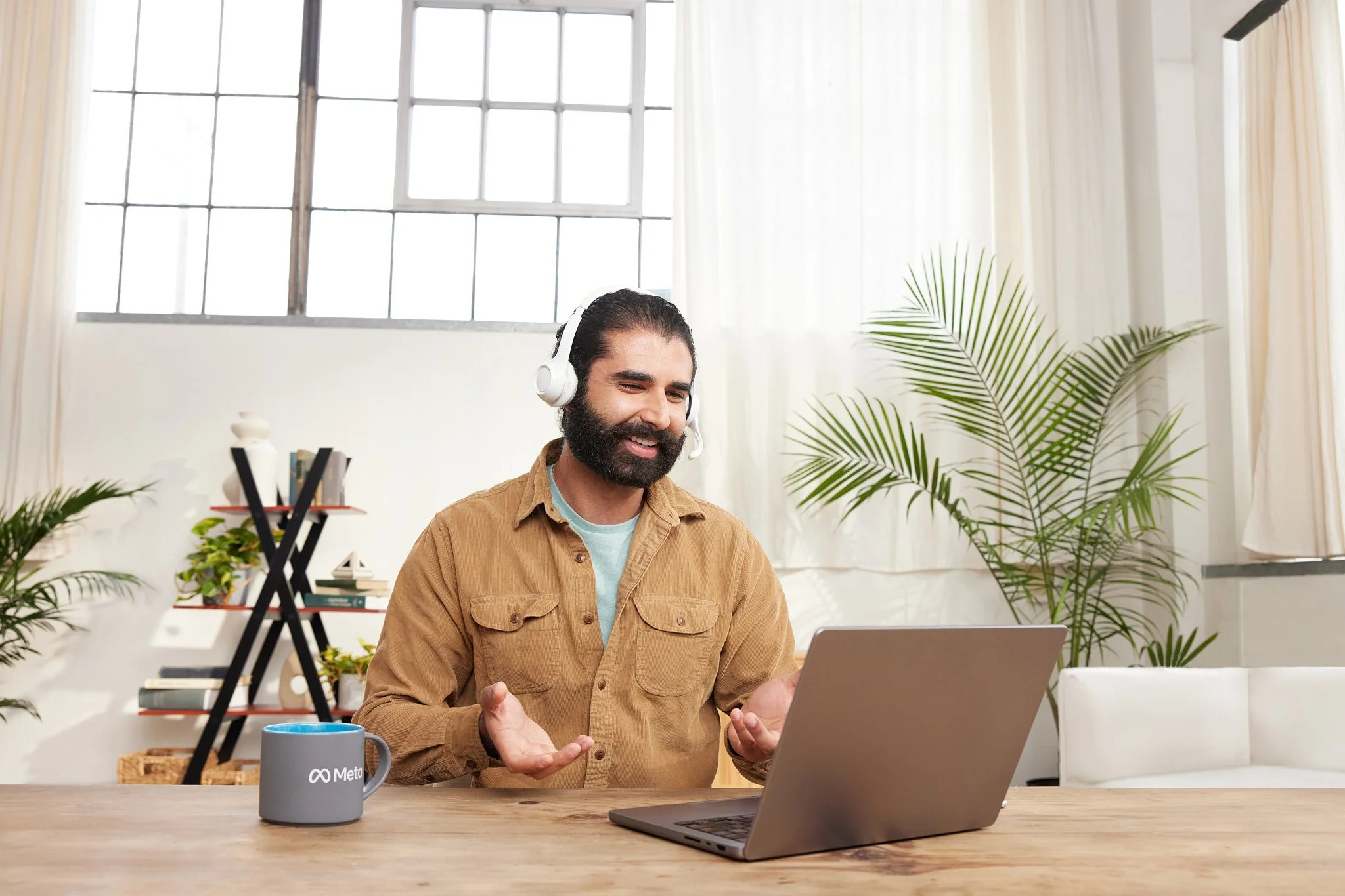 A man with a beard and long dark hair wearing a tan jacket and blue shirt, sitting at a desk with a laptop, wearing a headset, gesturing with his hands, in a bright room with large windows, plants, and books.