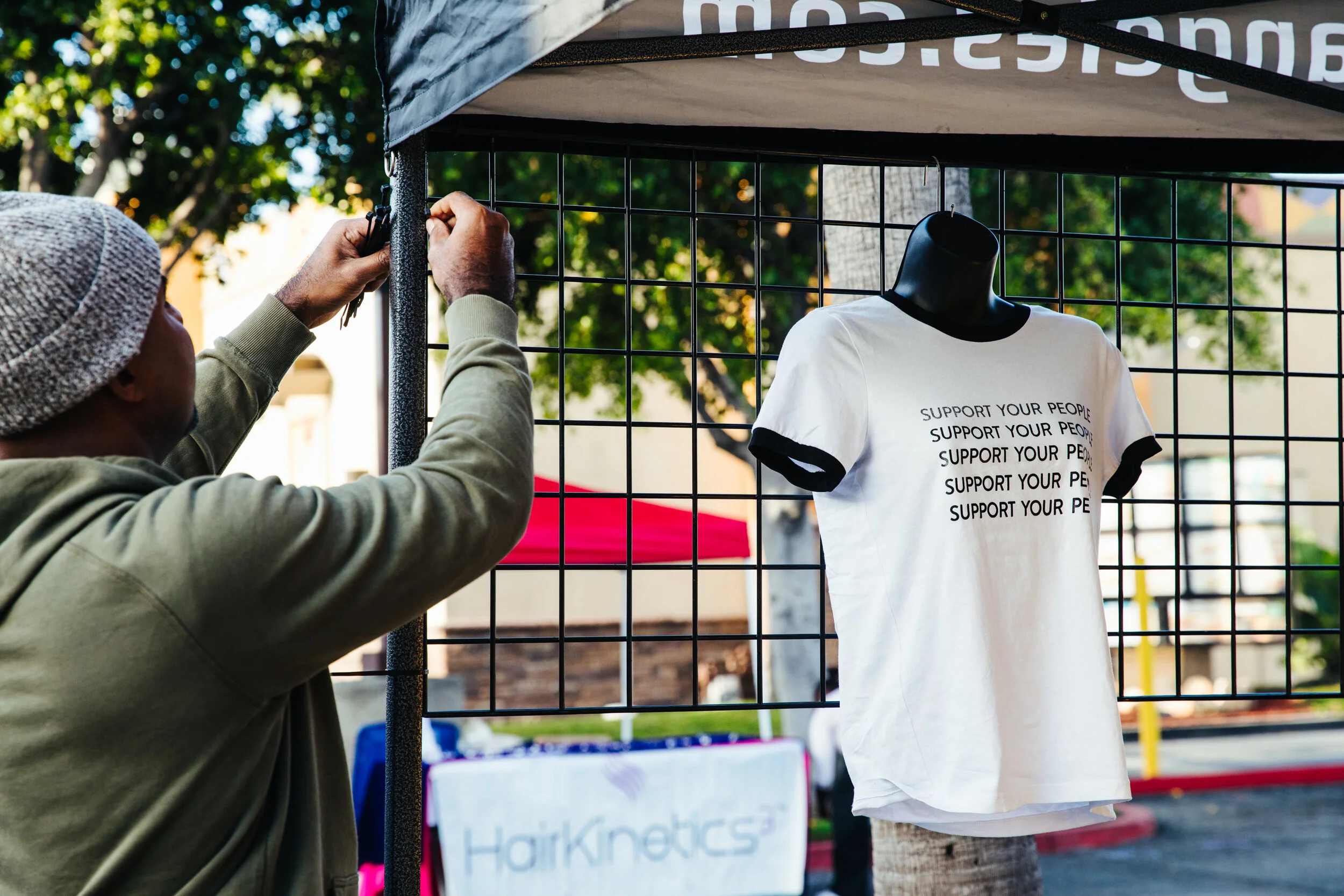 A person wearing a gray beanie and beige hoodie is securing a black metal grid fence with a clamp. Behind the fence, a white T-shirt with black trim and the printed message 'SUPPORT YOUR PEOPLE' hangs on a mannequin or display. The scene is outdoors, with trees and other booths or displays visible in the background.