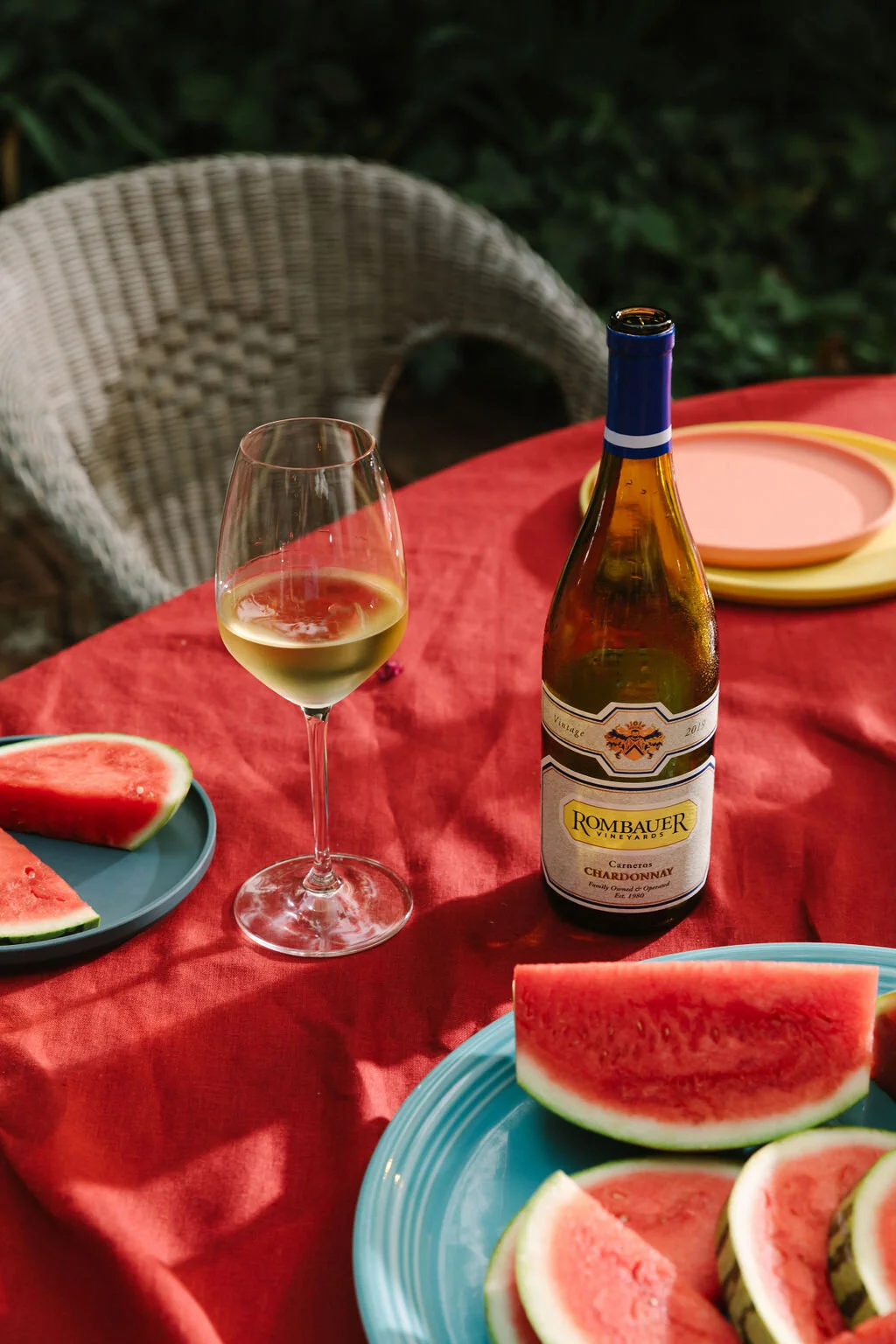 A glass of white wine, a bottle of Rombauer Chardonnay, slices of watermelon on plates, and colorful plates on a red tablecloth outdoors.