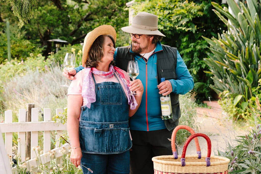 A man and a woman smiling and holding wine glasses in a garden, with the man holding a bottle of white wine and the woman in a sun hat and apron.
