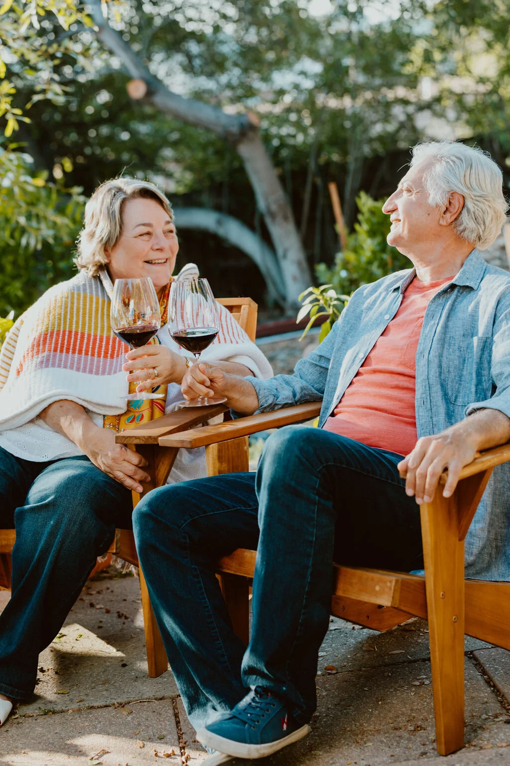 An elderly man and woman enjoying outdoor time with glasses of red wine, sitting on wooden chairs in a garden.