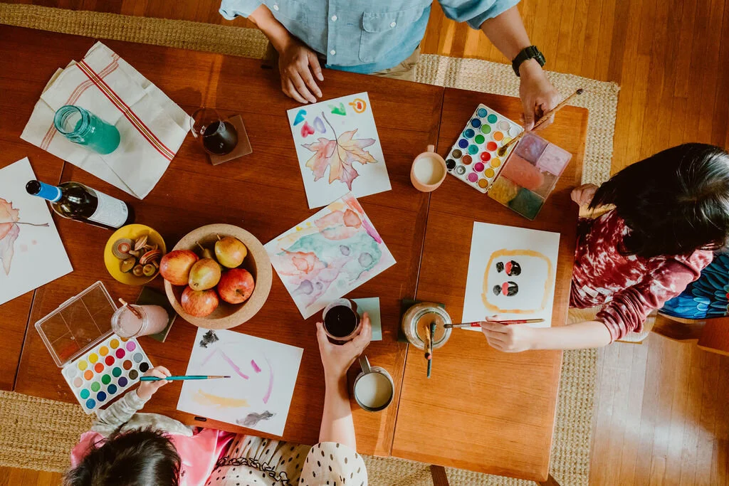 A group of children engaged in a watercolor painting activity at a wooden table with art supplies, including watercolors, paintbrushes, and paper, surrounded by apples and drinks.
