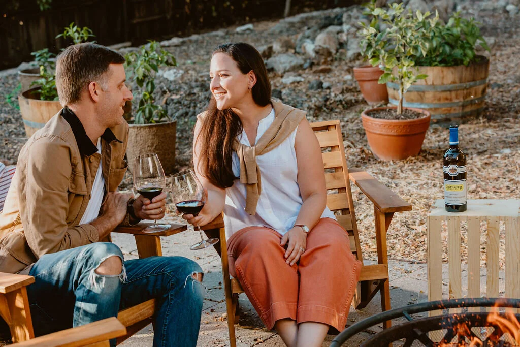 A man and woman sitting outside on wooden chairs, holding glasses of red wine and smiling at each other, with potted plants and a bottle of wine on a small table nearby.