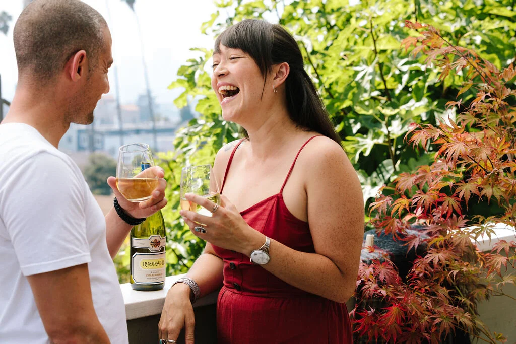 A man and woman are laughing and enjoying drinks on a balcony or patio surrounded by green and red foliage.