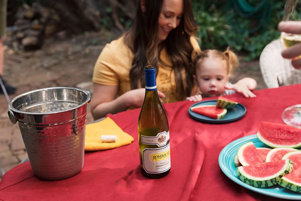 A woman and a young girl are sitting at a table outdoors with slices of watermelon and a bottle of white wine, surrounded by trees and greenery.