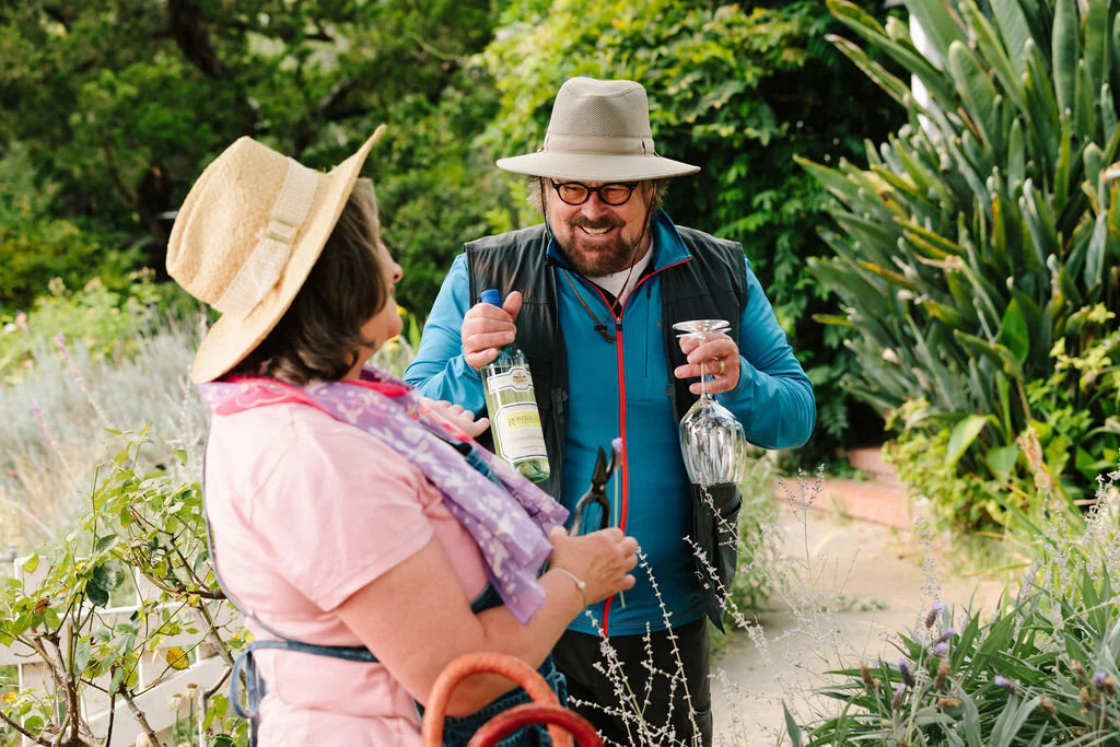 Two people, a man and a woman, are in a lush garden. The man is smiling, wearing a wide-brimmed hat, glasses, a blue jacket, and a black vest, and holding a wine glass and a bottle. The woman is wearing a straw hat, pink shirt, and a purple scarf, an