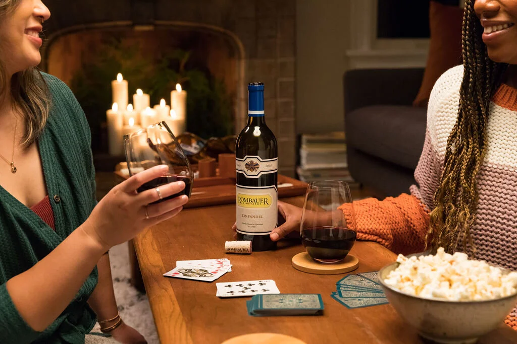 Two women playing cards and drinking wine at a cozy wooden table with lit candles in the background.