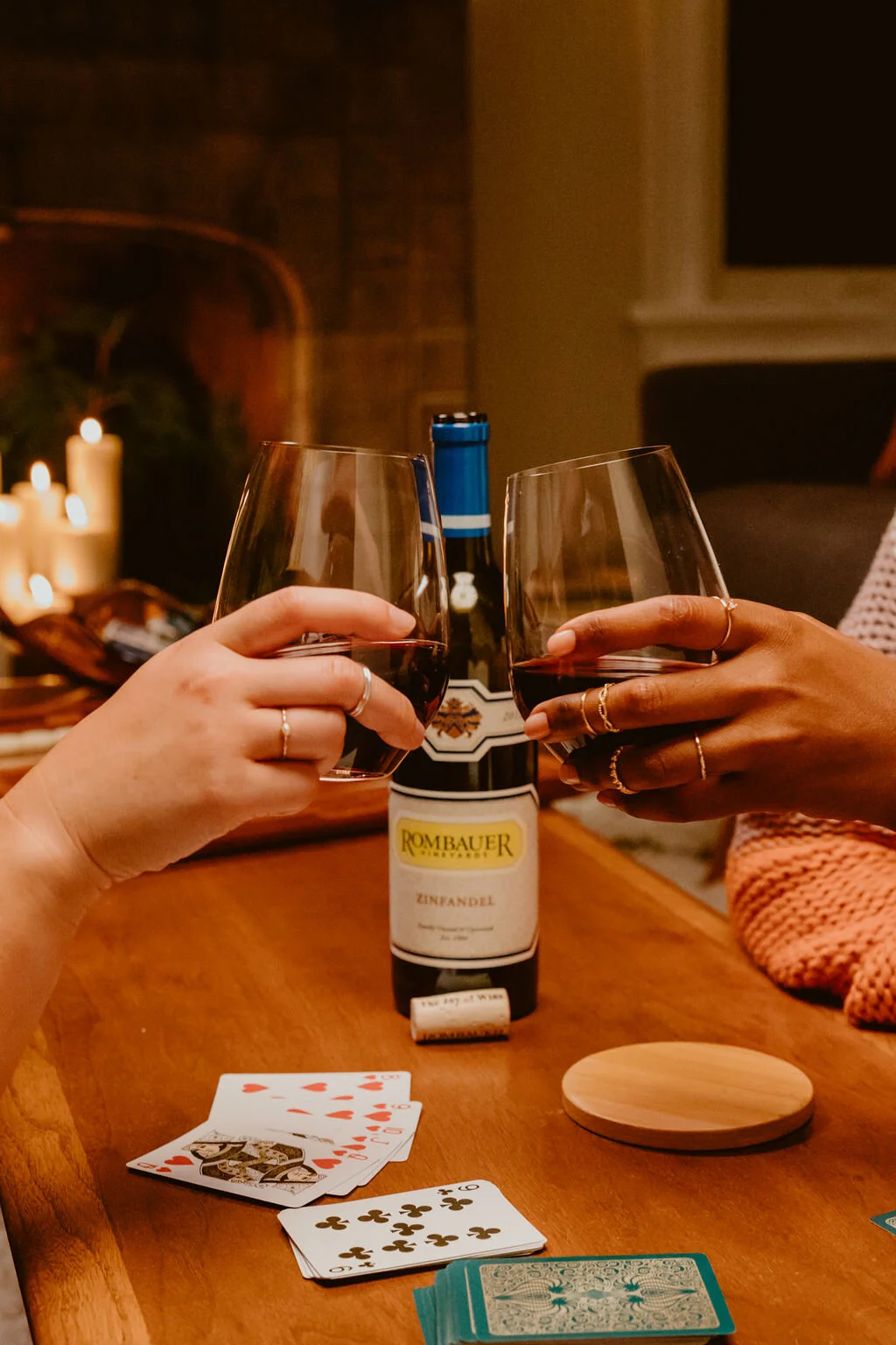 Two people clinking glasses of red wine in a cozy, indoor setting with a deck of playing cards on a wooden table, a wrapped wine bottle, and soft candlelight in the background.