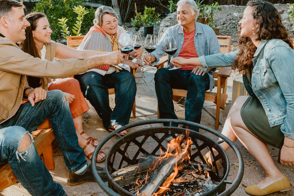 Group of six people sitting around a fire pit outdoors, clinking glasses of red wine, enjoying a social gathering.
