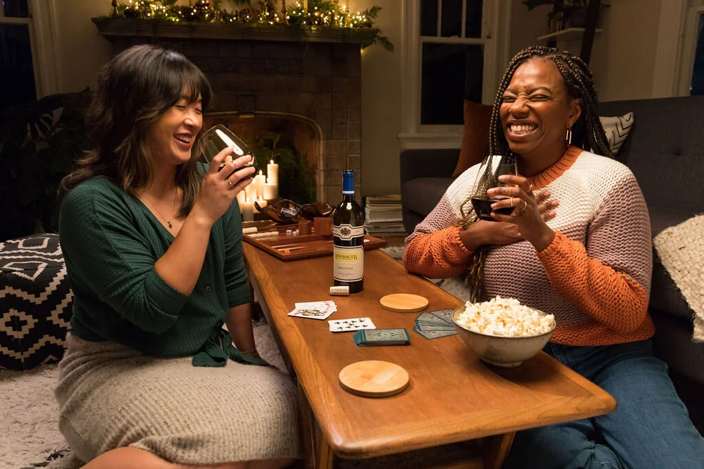 Two women are sitting at a wooden table, laughing and enjoying wine and popcorn, with a cozy decorated fireplace in the background.