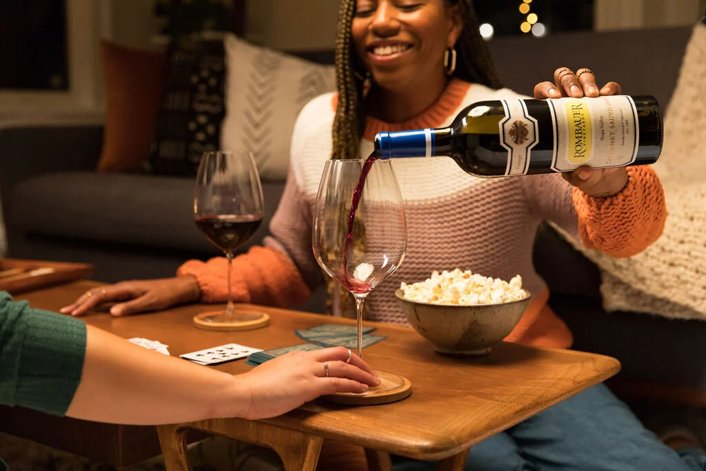 A woman is pouring red wine from a bottle into a glass on a wooden table, with a bowl of popcorn and a glass of wine nearby, while sitting with friends during a cozy gathering.