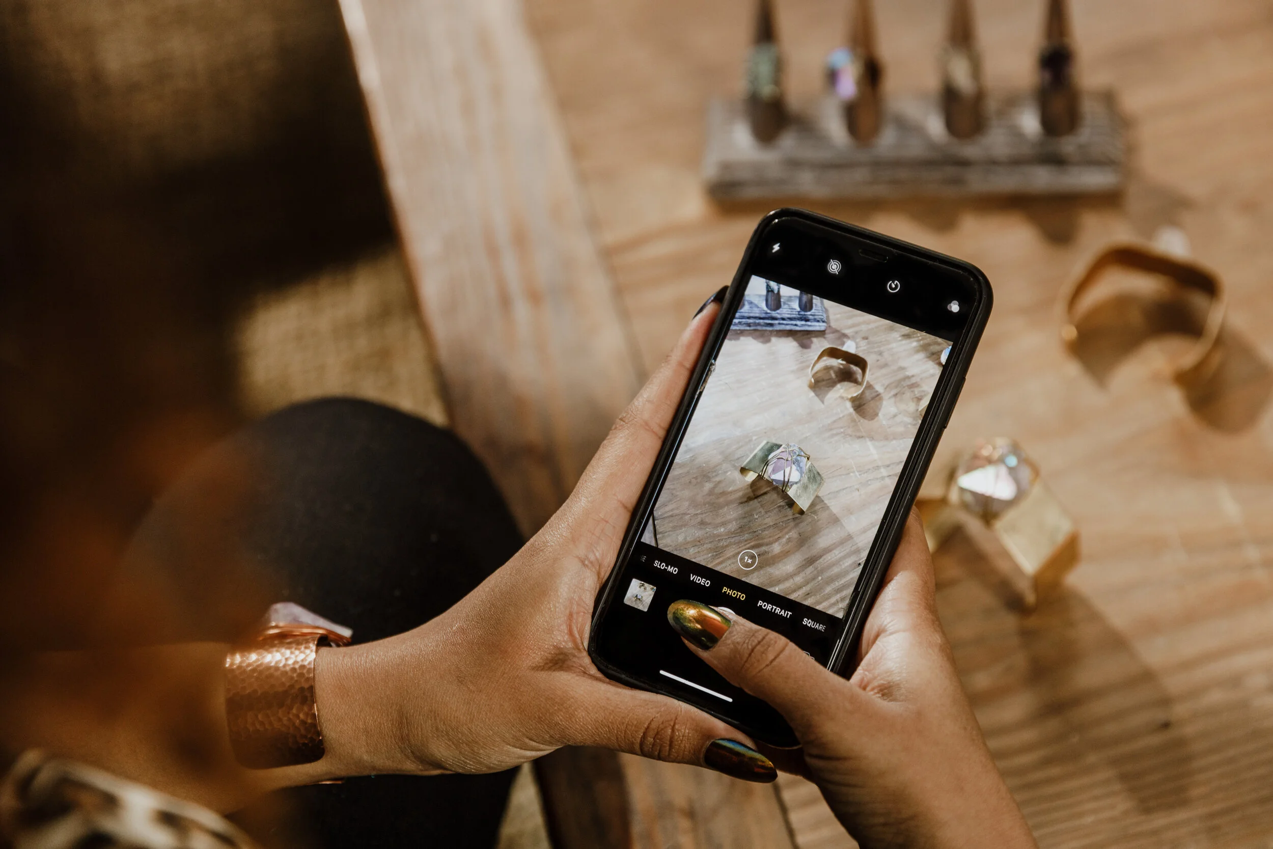 A person holding a smartphone taking a photo of jewelry on display on a wooden table.