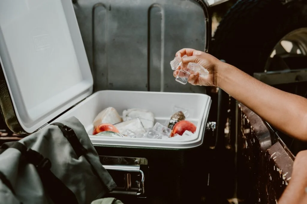 A person's hand is holding a crumpled ice cube above an open cooler filled with ice and various items, including apples and a bottle, outdoors near a vehicle.