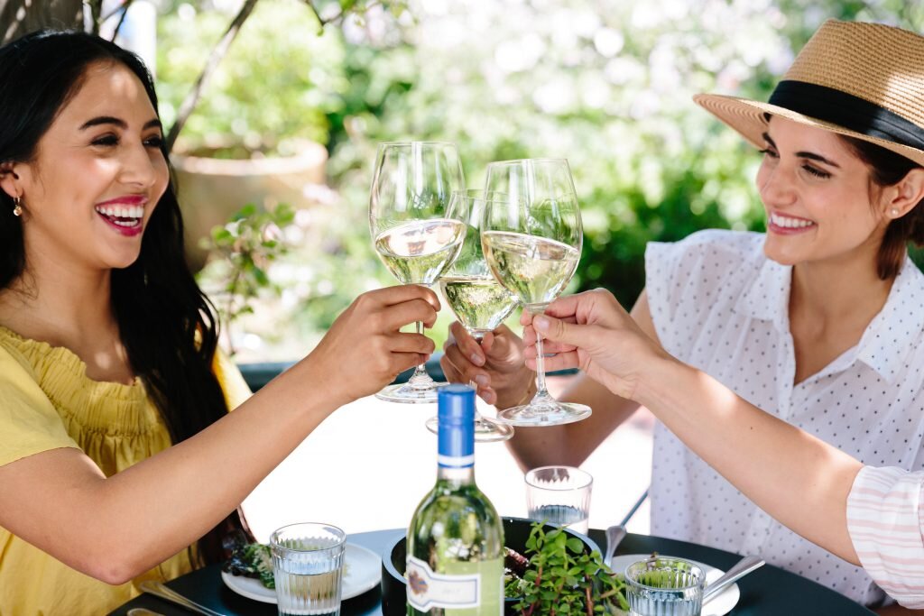 Two women clinking wine glasses together at an outdoor meal, smiling, with a bottle of white wine and a salad on the table.
