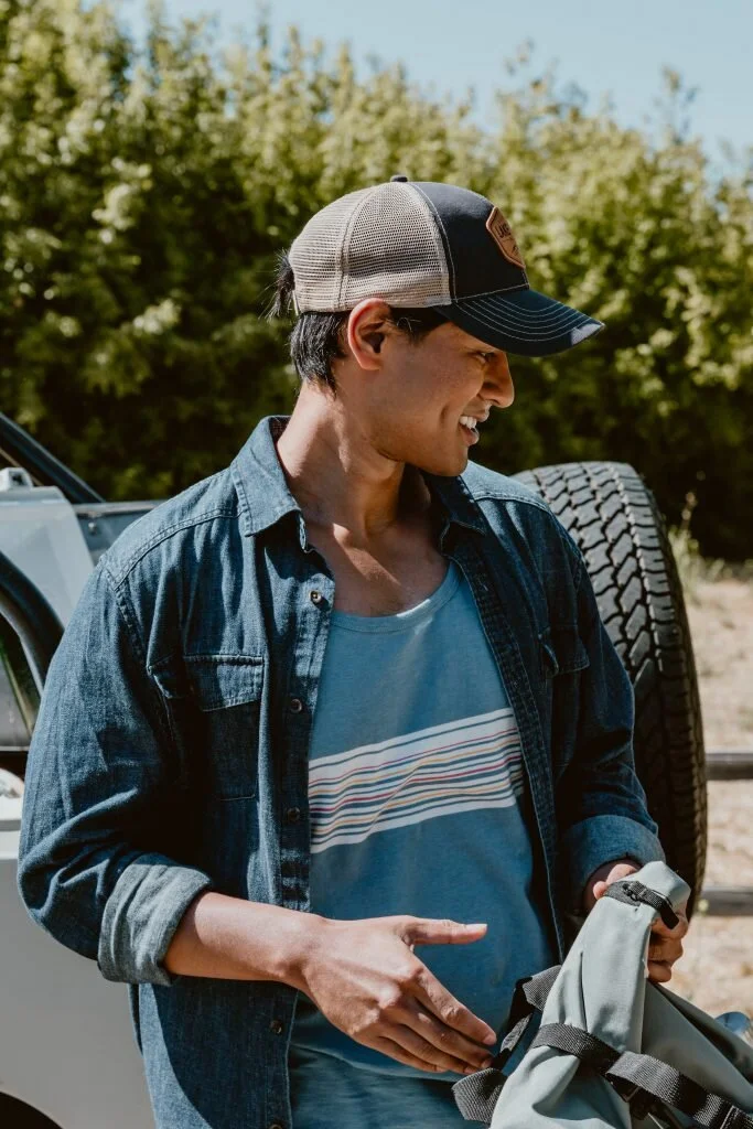 Young man smiling outdoors with a backpack, wearing a denim shirt, a striped t-shirt, and a trucker hat.