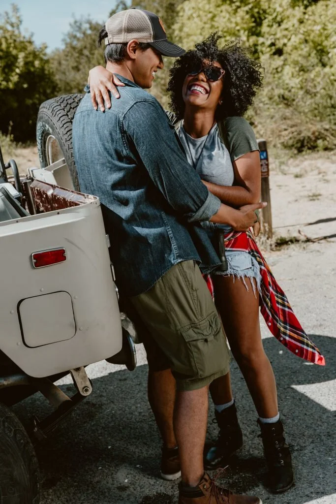 A smiling couple standing near a vehicle outdoors with trees in the background.