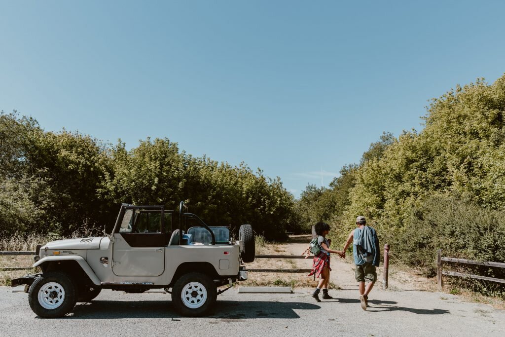 A couple holding hands and walking away from a parked outdoor vehicle on a rural dirt road, surrounded by trees and blue sky.