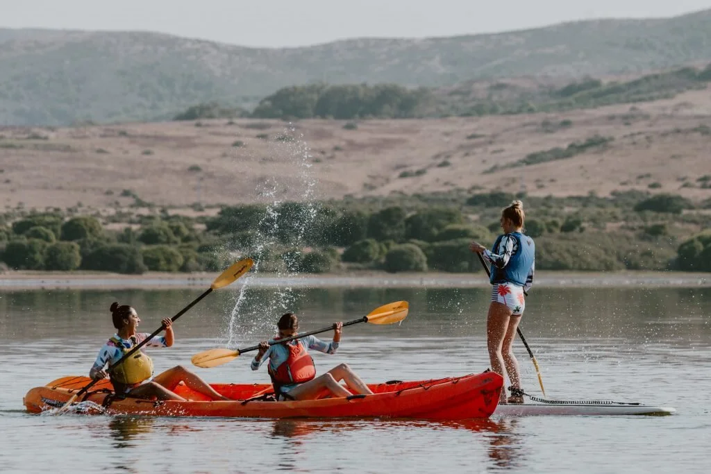 Three young women kayaking while another woman stands on a paddleboard taking notes on a clipboard on a calm lake with mountains in the background.