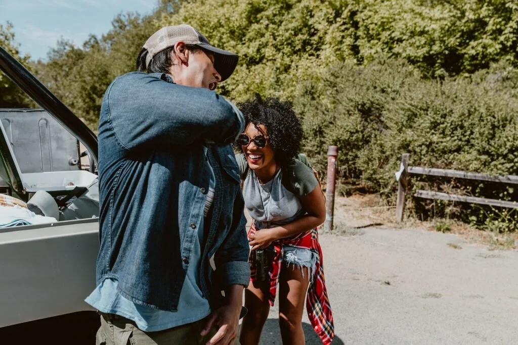 A young man and woman enjoy a moment outdoors with a forested background, laughing and smiling near a vehicle with its trunk open.