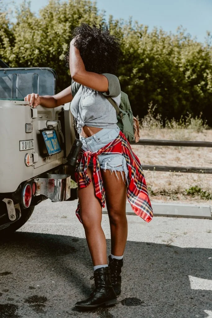 A woman with curly hair, wearing a gray t-shirt, denim shorts, a red plaid shirt tied around her waist, black combat boots, and a green backpack, standing next to a vintage vehicle on a road with a natural landscape background.