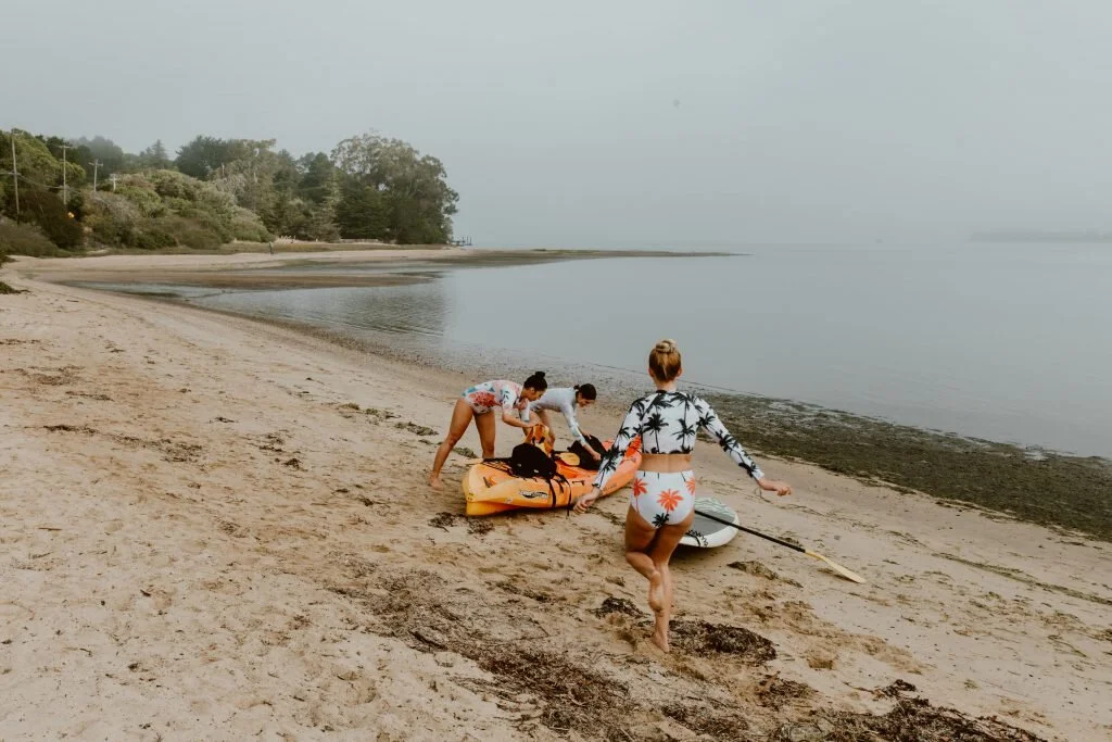 Three women at a beach preparing for water activities, with one walking towards the shore holding a paddleboard, and two others unpacking items from a kayak, during daytime with overcast sky.