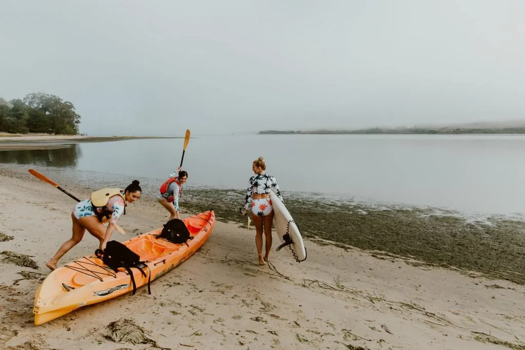 Three women preparing kayaks on a beach by a calm body of water on a cloudy day.
