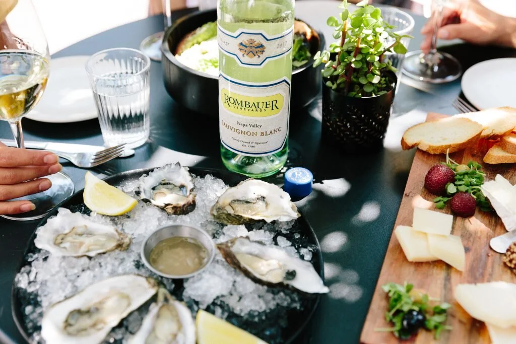 Oysters on a bed of crushed ice with lemon wedges, a bottle of white wine, a glass of white wine, and a charcuterie board with cheese, strawberries, and bread, on a dining table.