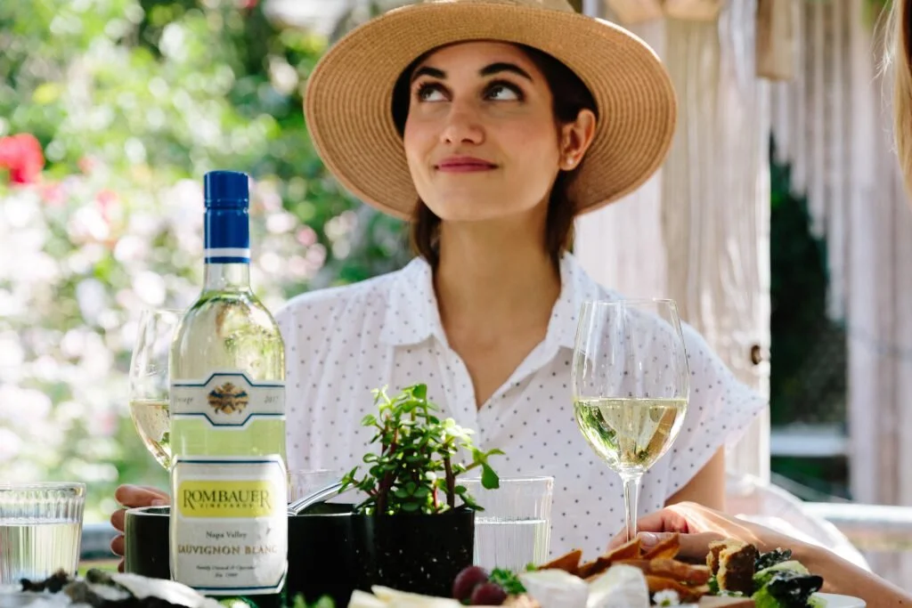 A woman wearing a large sun hat and a white polka dot shirt sitting outdoors at a table with a bottle of white wine, two wine glasses, a plant, and some food, with greenery in the background.