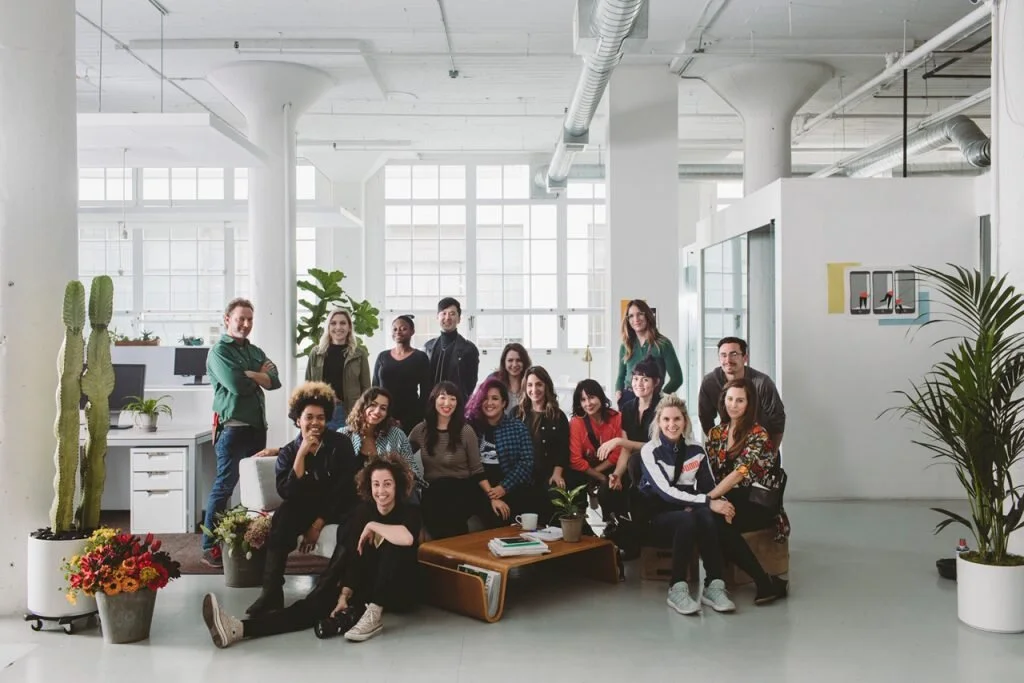 Group of diverse young adults smiling and posing in a modern bright office with large windows, plants, and casual seating.