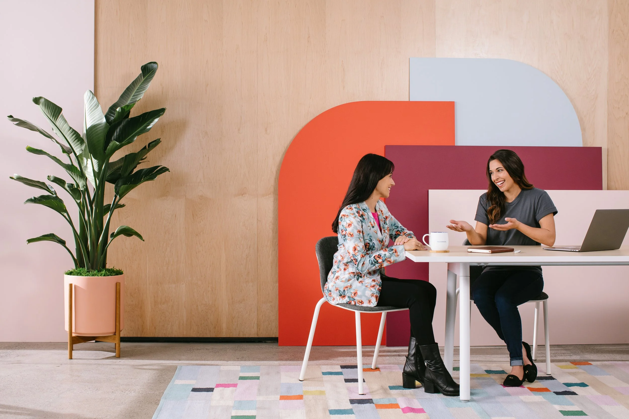 Two women sitting at a white table having a conversation in a modern office space. The woman on the left is wearing a floral blazer, and the woman on the right is wearing a grey top and black boots. A large green plant in a pink pot is on the left, and geometric colored panels are on the wall behind them.