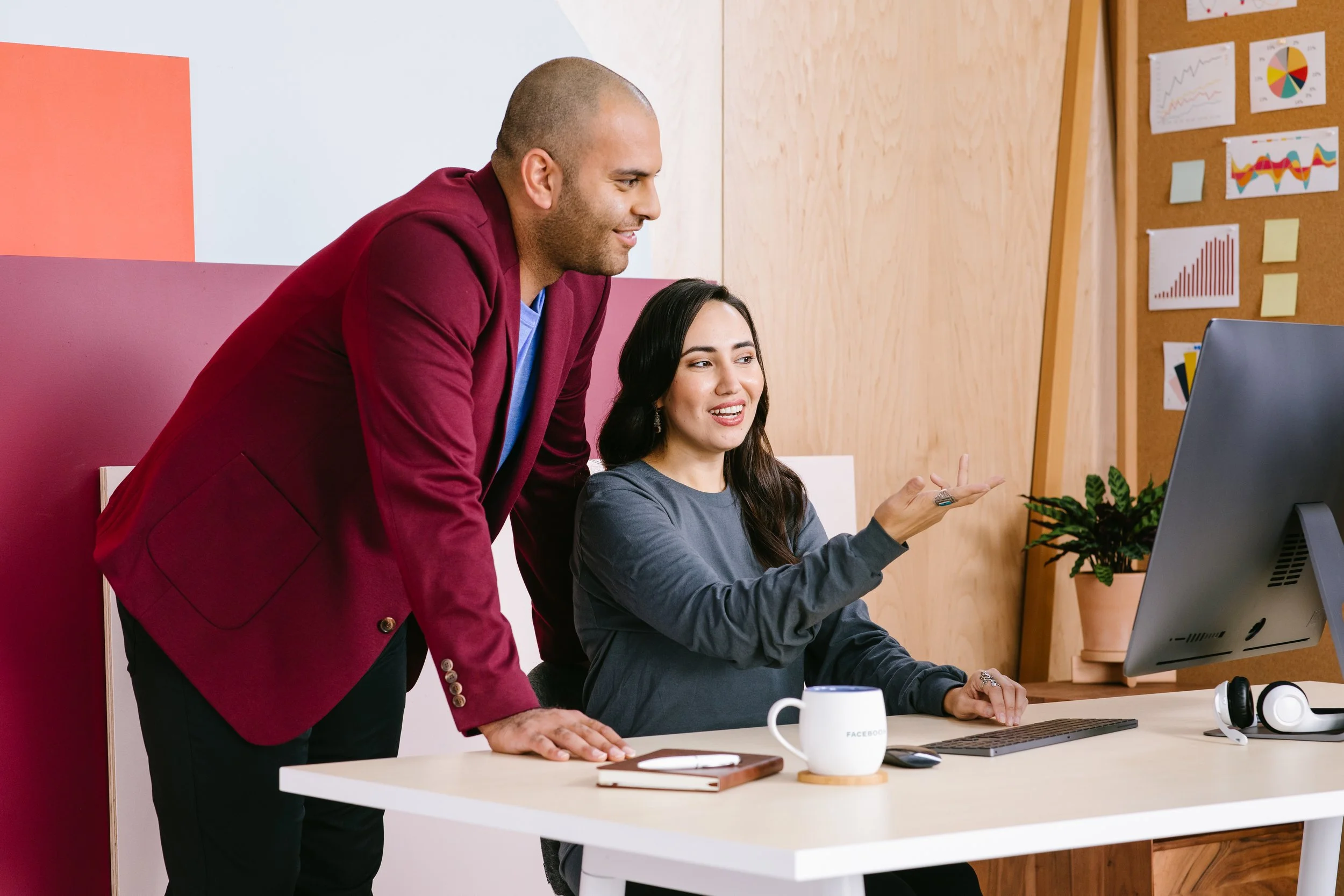A man in a red blazer leaning over a woman's shoulder as she sits at a desk in front of a computer in an office.
